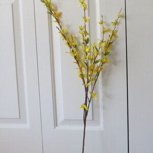 May include: A tall, artificial forsythia stem with vibrant yellow flowers and small green leaves. The stem is brown and slender, with multiple branches extending upwards. The flowers are clustered along the branches, creating a cheerful and natural look.