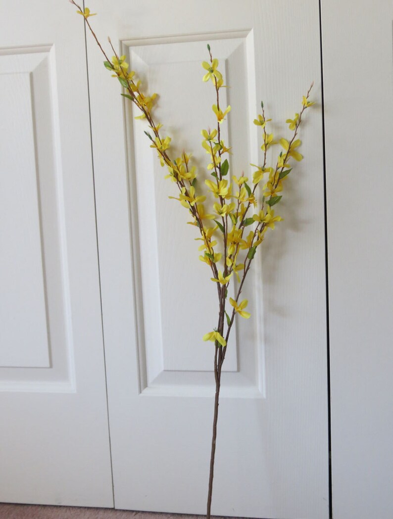 May include: A tall, artificial forsythia stem with vibrant yellow flowers and small green leaves. The stem is brown and slender, with multiple branches extending upwards. The flowers are clustered along the branches, creating a cheerful and natural look.