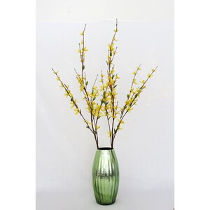 May include: A decorative arrangement featuring yellow forsythia branches in a green, ribbed glass vase. The vase has a metallic finish, and the branches are full of small yellow flowers and green leaves, creating a vibrant display.