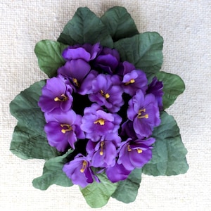 May include: A cluster of artificial African violet flowers featuring vibrant purple petals with small yellow centers, surrounded by a dense arrangement of textured, dark green leaves with scalloped edges, viewed from directly above.