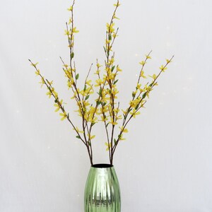 May include: A decorative arrangement featuring yellow forsythia branches in a green, ribbed glass vase. The vase has a metallic finish and a dark interior, creating a striking contrast with the bright flowers.