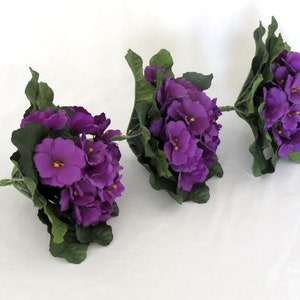 May include: Three artificial violet flower stems with vibrant purple blooms and green leaves. Each flower cluster has a central yellow stamen. The stems are a light green color. The flowers are arranged on a white background.