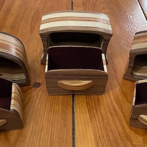 May include: Wooden jewelry boxes with a curved design. The boxes are made of layered wood in shades of brown and tan. Each box has a drawer with a burgundy interior. The boxes are on a wooden surface.