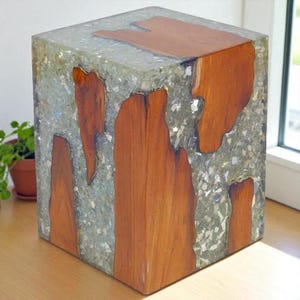 Peut inclure: Une table d'appoint carrée en bois et résine. Le bois, de couleur marron chaud, est intégré dans la conception, créant un effet visuel unique. La résine claire, parsemée de particules, entoure le bois. La table est de forme cubique.