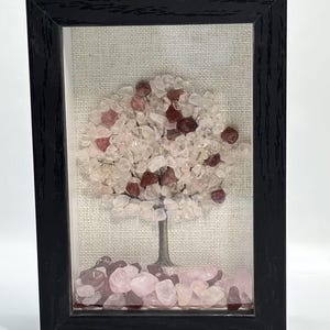 Pode incluir: Uma obra de arte emoldurada com um design de &aacute;rvore feito de pequenas pedras coloridas. A &aacute;rvore tem um tronco marrom e uma copa de pedras rosa claro e transparentes, com pedras vermelhas mais escuras espalhadas. A moldura &eacute; preta e o fundo &eacute; de tecido neutro.