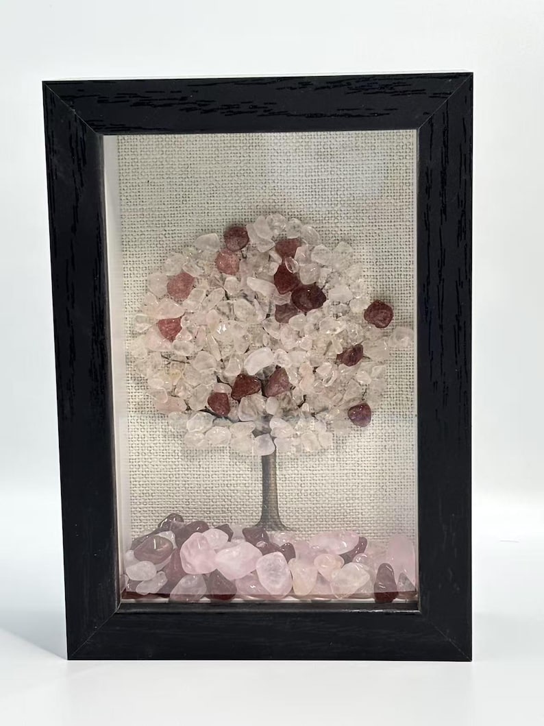 Pode incluir: Uma obra de arte emoldurada com um design de &aacute;rvore feito de pequenas pedras coloridas. A &aacute;rvore tem um tronco marrom e uma copa de pedras rosa claro e transparentes, com pedras vermelhas mais escuras espalhadas. A moldura &eacute; preta e o fundo &eacute; de tecido neutro.
