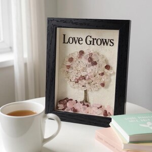 Pode incluir: Uma obra de arte emoldurada com o texto "Love Grows" acima de um desenho de &aacute;rvore feito de pedras rosa e transparentes. A moldura preta est&aacute; em uma superf&iacute;cie branca ao lado de uma caneca branca e livros.