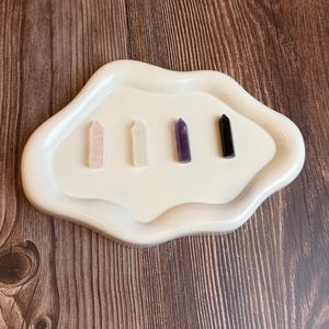 May include: A white, cloud-shaped tray holds four crystal points: rose quartz, clear quartz, amethyst, and black obsidian. The crystals are arranged in a row on the off-white tray, set against a wooden background.
