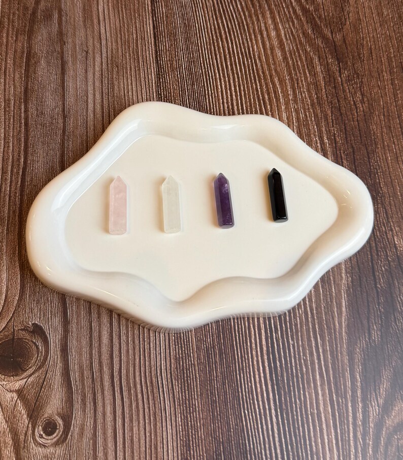 May include: A white, cloud-shaped tray holds four crystal points: rose quartz, clear quartz, amethyst, and black obsidian. The crystals are arranged in a row on the off-white tray, set against a wooden background.