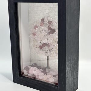 Pode incluir: Uma escultura decorativa de &aacute;rvore emoldurada. A &aacute;rvore apresenta um tronco de metal escuro e uma coroa arredondada feita de pedras rosa claro e transparentes. A base da &aacute;rvore &eacute; cercada por pedras rosa, tudo emoldurado em uma moldura de madeira preta.
