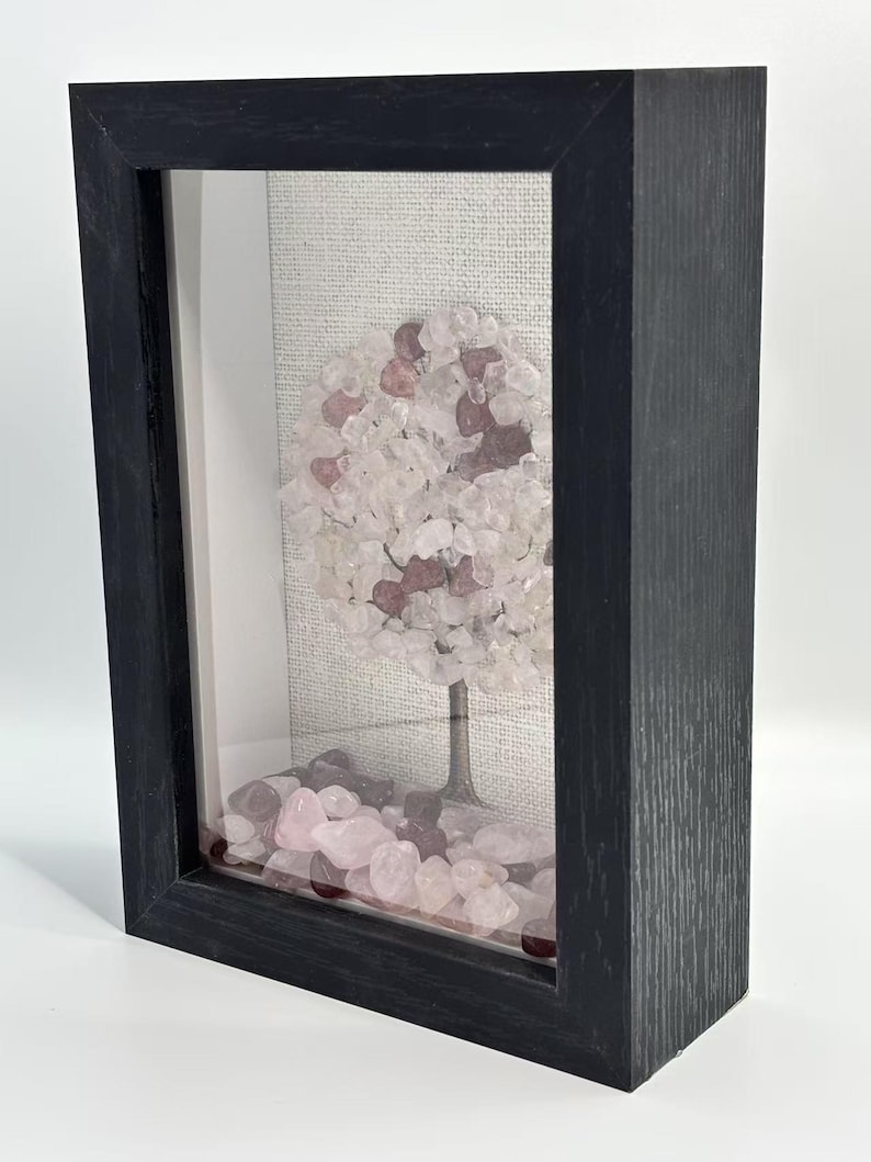 Pode incluir: Uma escultura decorativa de &aacute;rvore emoldurada. A &aacute;rvore apresenta um tronco de metal escuro e uma coroa arredondada feita de pedras rosa claro e transparentes. A base da &aacute;rvore &eacute; cercada por pedras rosa, tudo emoldurado em uma moldura de madeira preta.