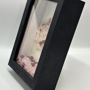 Pode incluir: Uma caixa de sombra de madeira preta com frente de vidro, inclinada para a direita. No interior, pedras preciosas rosa e roxas s&atilde;o dispostas contra um fundo de tecido de cor clara. A moldura tem uma apar&ecirc;ncia de gr&atilde;o de madeira texturizada.