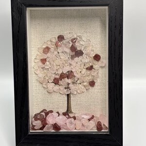 Pode incluir: Uma obra de arte emoldurada com uma &aacute;rvore feita de pedras preciosas rosa e claras. A &aacute;rvore tem um tronco marrom e uma copa de pedras rosa claro e vermelhas, contra um fundo de tecido neutro. A moldura &eacute; preta.