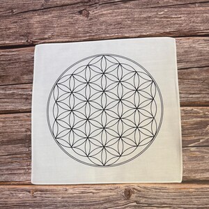 May include: Square white decorative item with a black geometric flower of life design. The design is a symmetrical pattern of overlapping circles. The item is on a wooden surface.