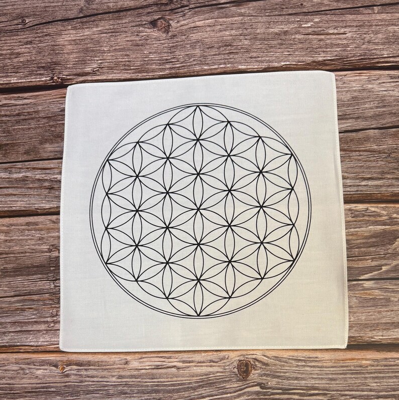 May include: Square white decorative item with a black geometric flower of life design. The design is a symmetrical pattern of overlapping circles. The item is on a wooden surface.