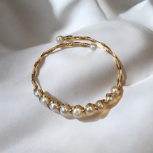 May include: A gold-toned bracelet featuring a twisted wire design. The bracelet is adorned with several white pearls, and the ends of the bracelet have two additional pearls. The jewelry is displayed on a white, silky surface.