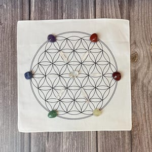 May include: A white square cloth with a black Flower of Life design, surrounded by seven colorful gemstones and clear quartz crystals. The gemstones are purple, red, blue, green, yellow, and clear.