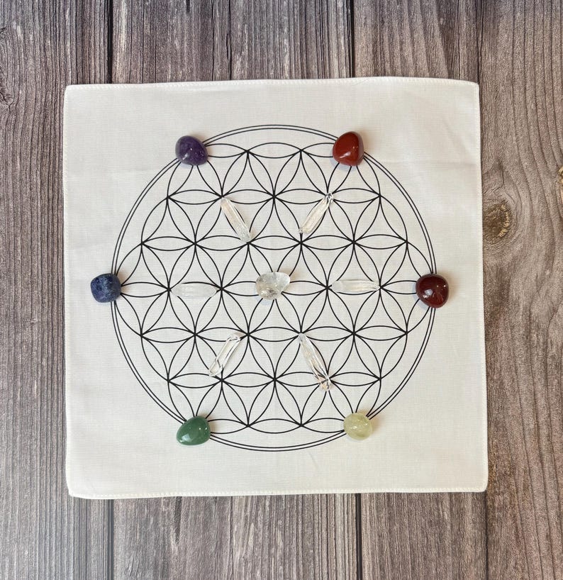 May include: A white square cloth with a black Flower of Life design, surrounded by seven colorful gemstones and clear quartz crystals. The gemstones are purple, red, blue, green, yellow, and clear.