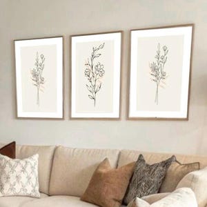 Può includere: Tre stampe artistiche botaniche incorniciate. Ogni stampa presenta un delicato disegno a tratto nero di fiori e fogliame su sfondo crema. Le cornici sono in una calda tonalità legno. Le stampe sono esposte sopra un divano beige con cuscini decorativi.