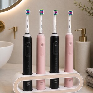Könnte beinhalten: Vier elektrische Zahnbürsten, zwei schwarze und zwei rosafarbene, stehen in einem ovalen Halter in Weiß und heller Holzoptik. Jede Zahnbürste hat einen weißen Kopf mit bunten Borsten und das Oral-B Braun Logo am Griffende.