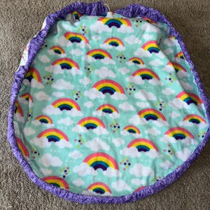 May include: A round, light blue blanket with a repeating pattern of rainbows and white clouds. The blanket has a purple ruffled edge. The rainbows are in shades of red, orange, yellow, green, blue, and purple.