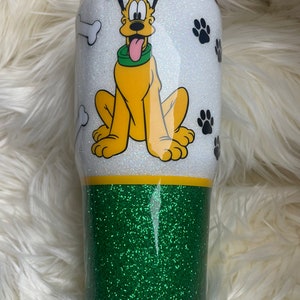 May include: A white and green glitter tumbler with a cartoon image of Pluto the dog. The tumbler has a white top with a black outline and a green bottom. The image of Pluto is in color and shows him sitting with his tongue sticking out. There are paw prints and bone shapes around Pluto.