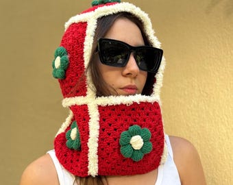 May include: A red and white crocheted hood with green and white flower accents. The hood is worn with black sunglasses and a white tank top. The hood is a unique fashion accessory.