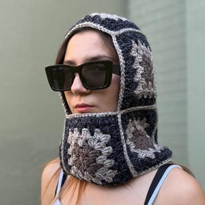 May include: A handmade crochet balaclava in a patchwork design. The balaclava features a repeating square pattern in shades of gray, brown, and beige. The head covering is worn with black sunglasses and a light blue tank top.