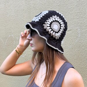 Op de afbeelding: Een zwarte gehaakte vissershoed met een bloemenontwerp in grijstinten en wit. De hoed heeft een geschulpte rand. De persoon draagt een grijze tanktop.