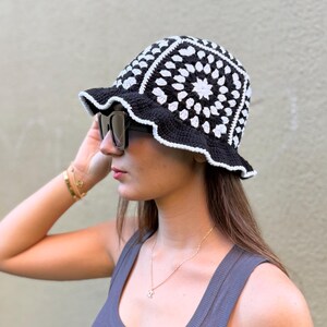 May include: A black and white crochet bucket hat with a scalloped brim. The hat features a geometric pattern with white squares and a black border. The hat is worn by a person wearing sunglasses and a gray tank top.