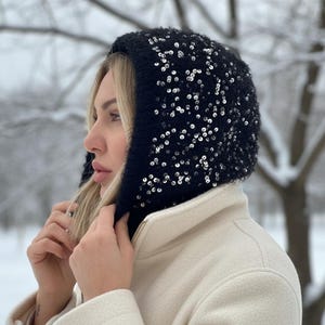 May include: A black knit hood with silver sequins, worn over a cream-colored coat. The hood is a winter accessory, offering warmth and style. The background shows a snowy, outdoor scene.