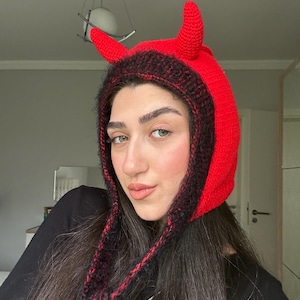 Puede incluir: Un gorro de cuernos de diablo tejido en rojo y negro. El gorro presenta dos cuernos rojos y un ribete negro y rojo alrededor de la cara. El gorro tiene lazos largos.
