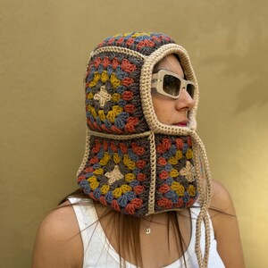 May include: A colorful crochet hood with a granny square design. The hood features a mix of blue, yellow, red, and tan squares, outlined in a darker brown. The hood has a tan trim and ties, and is worn with sunglasses.