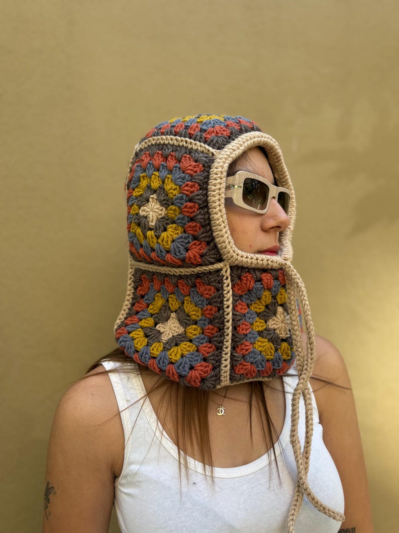 May include: A colorful crochet hood with a granny square design. The hood features a mix of blue, yellow, red, and tan squares, outlined in a darker brown. The hood has a tan trim and ties, and is worn with sunglasses.