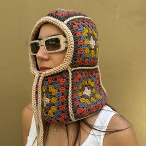 May include: A colorful crochet hood with a granny square design. The hood features a mix of brown, beige, yellow, blue, and orange squares. It has a beige trim and drawstrings. The person is wearing sunglasses and a white tank top.