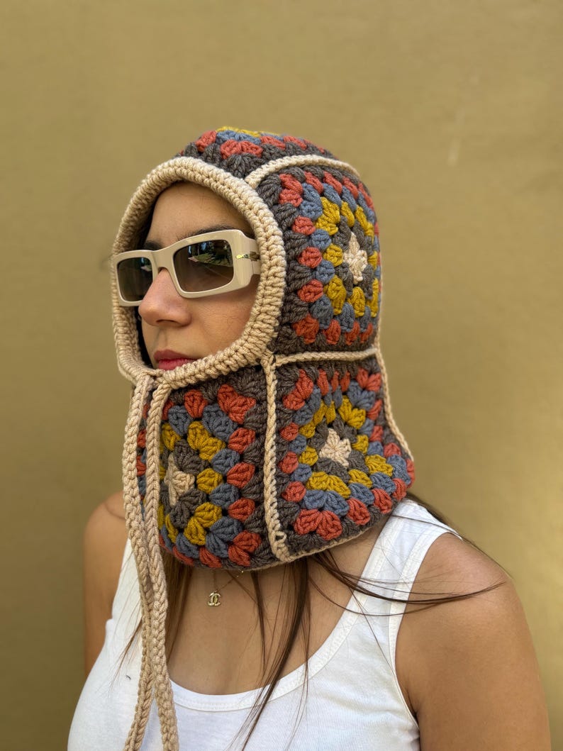 May include: A colorful crochet hood with a granny square design. The hood features a mix of brown, beige, yellow, blue, and orange squares. It has a beige trim and drawstrings. The person is wearing sunglasses and a white tank top.