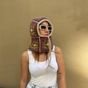 May include: A colorful crochet hood with a square pattern in shades of blue, red, yellow, and beige. The hood has a beige trim and ties, and is worn with sunglasses. The hood covers the head and neck.