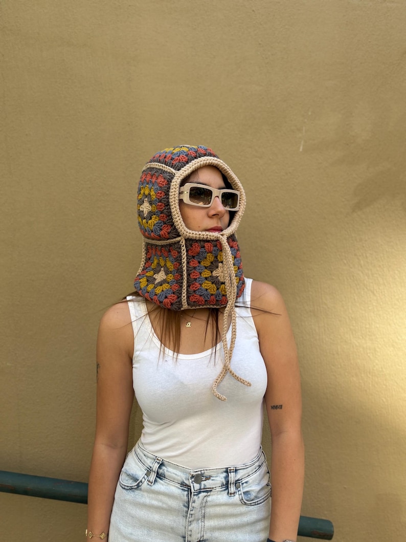 May include: A colorful crochet hood with a square pattern in shades of blue, red, yellow, and beige. The hood has a beige trim and ties, and is worn with sunglasses. The hood covers the head and neck.