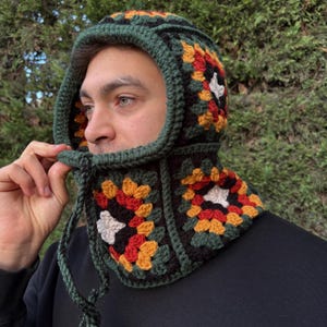 May include: A handmade crochet hood in a granny square pattern. The hood features a dark green trim and drawstring, with squares in black, red, orange, yellow, and white. The hood is designed to cover the head and neck.