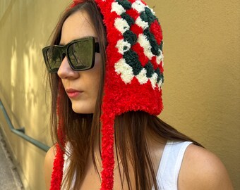 May include: A red, white, and green crocheted hat with long, red ties. The hat features a checkered pattern and is worn by a person wearing black sunglasses and a white tank top. The background is a yellow wall.