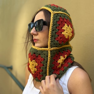 May include: A crocheted hood in a granny square pattern, featuring olive green, red, and yellow squares. The hood has a yellow border and is worn around the neck and head. The person is wearing black sunglasses and a white tank top.