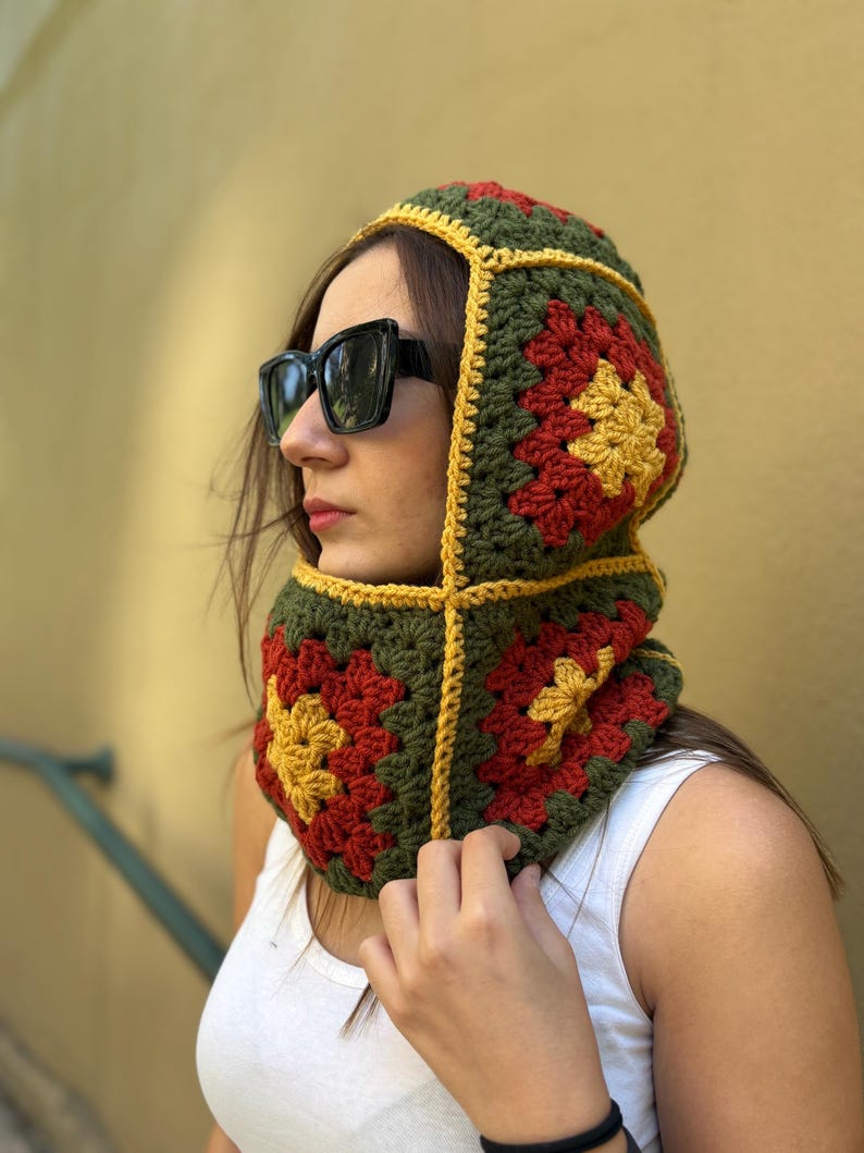May include: A crocheted hood in a granny square pattern, featuring olive green, red, and yellow squares. The hood has a yellow border and is worn around the neck and head. The person is wearing black sunglasses and a white tank top.