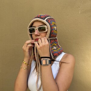 May include: A colorful crochet hood with a square pattern in shades of brown, yellow, blue, and red. The hood has a beige trim and long ties. The person is wearing beige sunglasses and a white tank top.