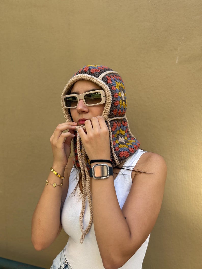 May include: A colorful crochet hood with a square pattern in shades of brown, yellow, blue, and red. The hood has a beige trim and long ties. The person is wearing beige sunglasses and a white tank top.