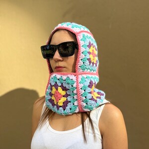 May include: A colorful crochet balaclava in a granny square pattern. The balaclava features a mix of pastel colors including pink, blue, yellow, and purple. The balaclava is worn with black sunglasses and a white tank top.