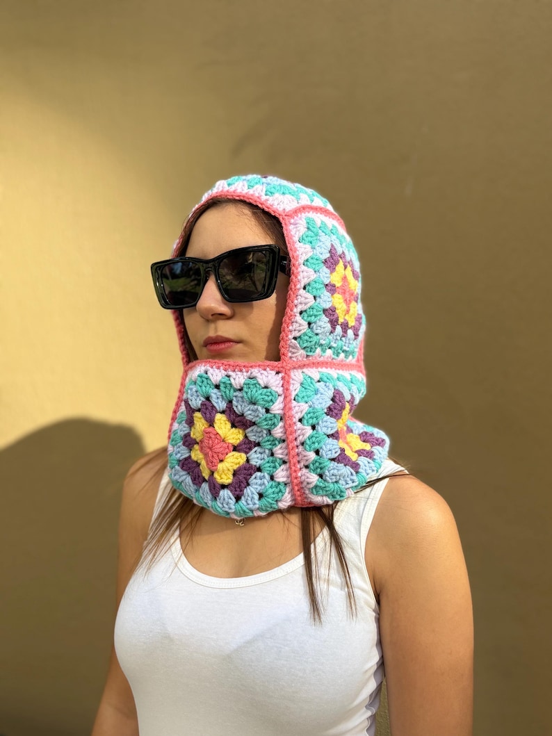 May include: A colorful crochet balaclava in a granny square pattern. The balaclava features a mix of pastel colors including pink, blue, yellow, and purple. The balaclava is worn with black sunglasses and a white tank top.