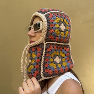 May include: A colorful crochet hood in a granny square pattern. The hood features squares of brown, orange, yellow, blue, and beige. The hood has a beige trim and drawstrings. The hood is worn with sunglasses.