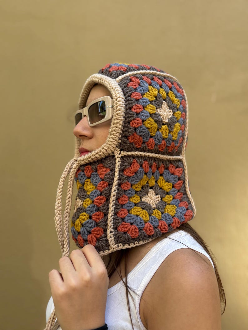 May include: A colorful crochet hood in a granny square pattern. The hood features squares of brown, orange, yellow, blue, and beige. The hood has a beige trim and drawstrings. The hood is worn with sunglasses.
