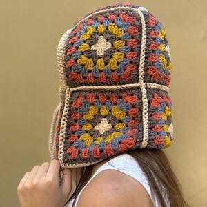May include: A colorful crochet hat with a square pattern design. The hat features a mix of colors including brown, yellow, blue, and orange. The hat has a beige trim and ties.