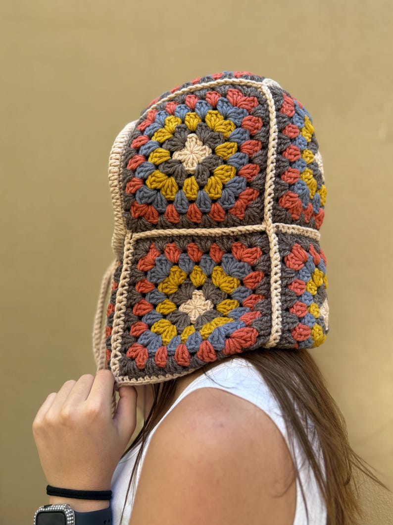 May include: A colorful crochet hat with a square pattern design. The hat features a mix of colors including brown, yellow, blue, and orange. The hat has a beige trim and ties.
